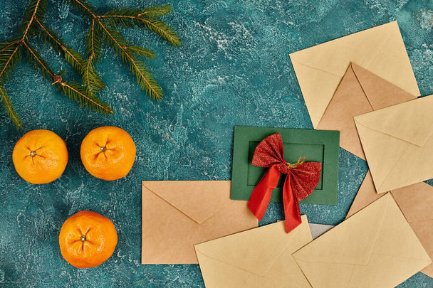 grüne Karte mit roter Schleife in der Nähe von Briefumschlägen, Mandarinen und Tannenzweigen auf blauem Hintergrund, Weihnachten - Foto, Bild