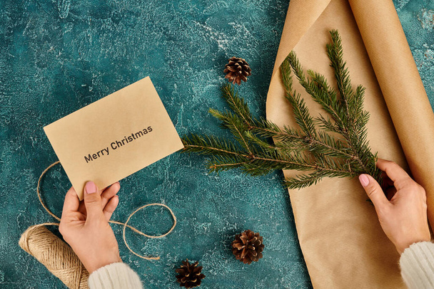 cropped view of woman with Merry Christmas envelope and pine branch near craft paper and twine, diy - Photo, Image