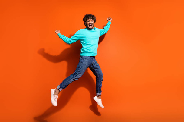 Full length photo of sportive indian guy curls hair wear denim jeans with cyan pullover win lottery isolated on orange color background. - Fotoğraf, Görsel