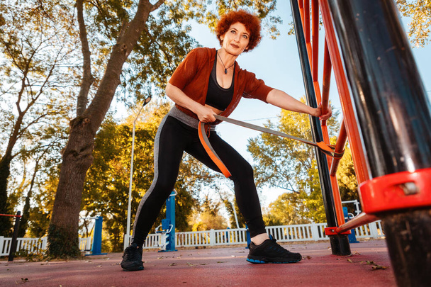 Visão inferior da mulher caucasiana forte adulta faz alongamento com banda elástica no playground esportivo. - Foto, Imagem
