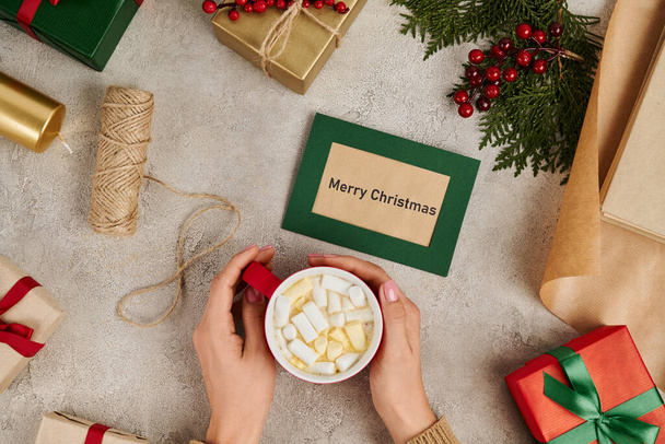 vista recortada de la mujer sosteniendo chocolate caliente con malvaviscos cerca de la tarjeta de felicitación de Feliz Navidad - Foto, Imagen
