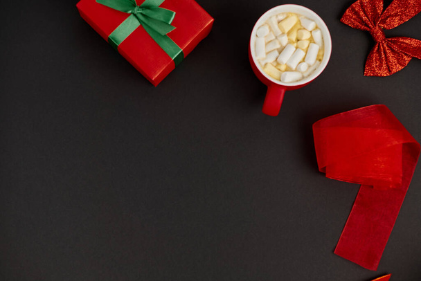 κούπα ζεστή σοκολάτα με ζαχαρωτό κοντά σε κουτί δώρου και γυαλιστερό φιόγκο με κόκκινη κορδέλα σε μαύρο φόντο - Φωτογραφία, εικόνα