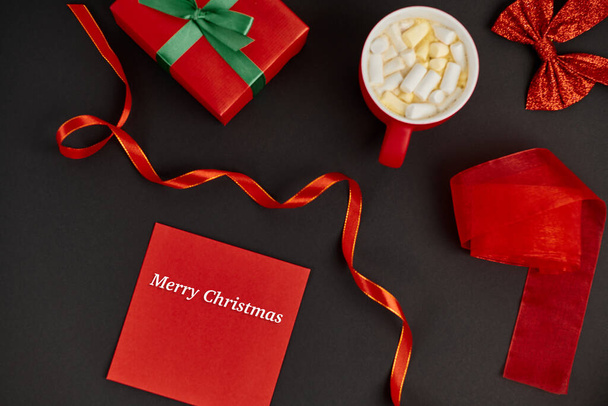 piros boríték boldog karácsonyi felirattal doboz közelében és forró csokoládé mályvacukorral fekete - Fotó, kép