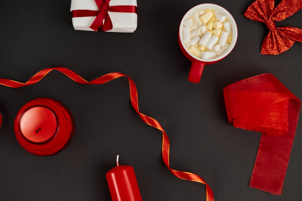 taza de chocolate caliente con malvavisco cerca de cintas rojas y velas en negro, fondo de Navidad - Foto, imagen
