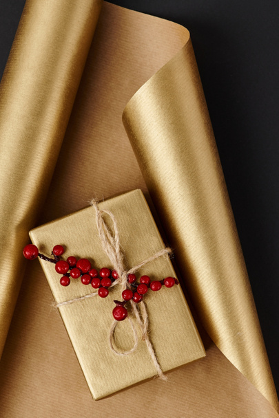 golden gift box with red holly berries on shiny wrapping paper and black backdrop, Christmas decor - Photo, Image