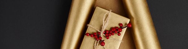 блестящая подарочная коробка с красными ягодами падуба на золотой бумаге и черном фоне, горизонтальный баннер - Фото, изображение