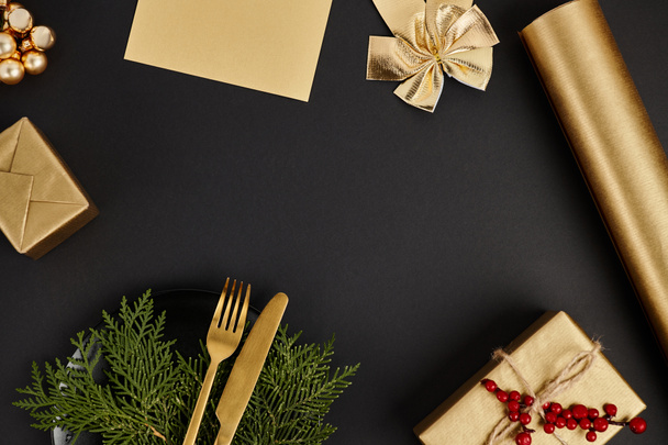 goldenes Besteck auf Wacholderzweigen in der Nähe von glänzendem Weihnachtsdekor auf schwarzem Hintergrund, festlicher Rahmen - Foto, Bild