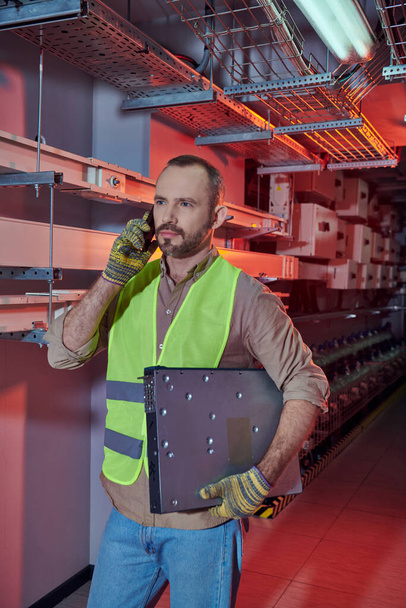 συγκεντρωμένος τεχνικός με γιλέκο ασφαλείας και γάντια που μιλάει τηλεφωνικώς και κοιτάζει αλλού κατά τη διάρκεια της εργασίας - Φωτογραφία, εικόνα