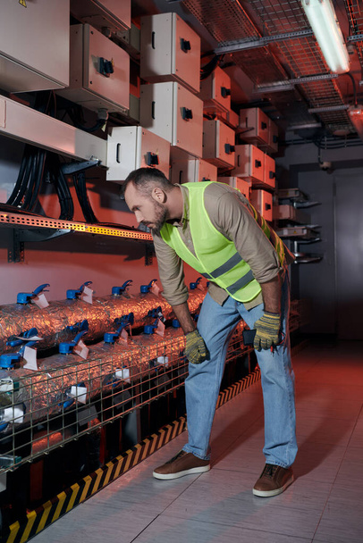 atractivo técnico concentrado en guantes de seguridad y chaleco inclinado mientras trabaja en el centro de datos - Foto, Imagen