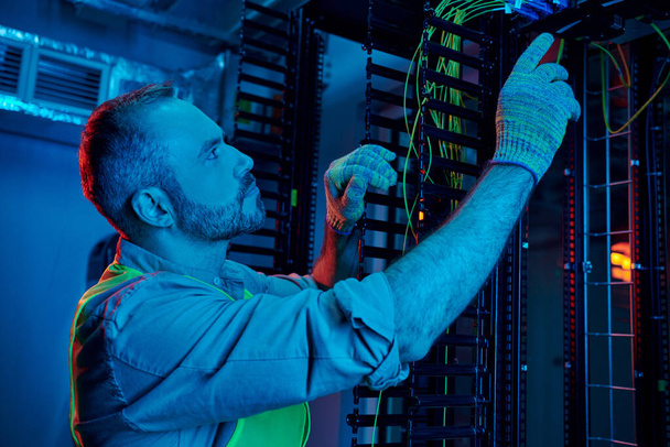 good looking concentrated technician in safety gloves and vest inspecting cables, data center - Photo, Image