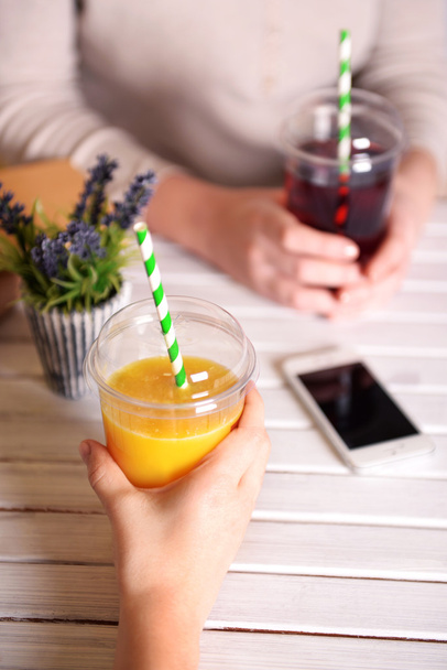 Dvě ženské ruce s fast food šálky šťávy a mobilní telefon na dřevěný stůl s rostlinou na světlé zdi pozadí - Fotografie, Obrázek