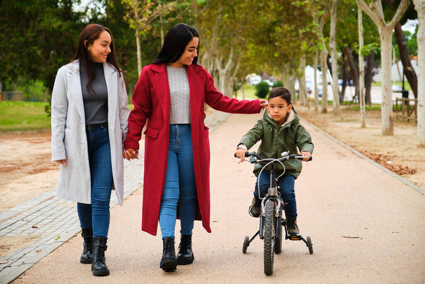Latin lesbian couple walking in a park with their son in a bicycle. LGBT family. - Photo, Image