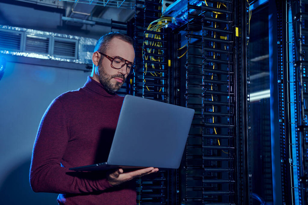 good looking pensive data center specialist in turtleneck with glasses and beard looking at laptop - Photo, Image