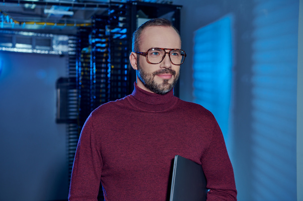 alegre centro de datos guapo especialista en cuello alto con gafas que sostienen el ordenador portátil y mirando hacia otro lado - Foto, Imagen