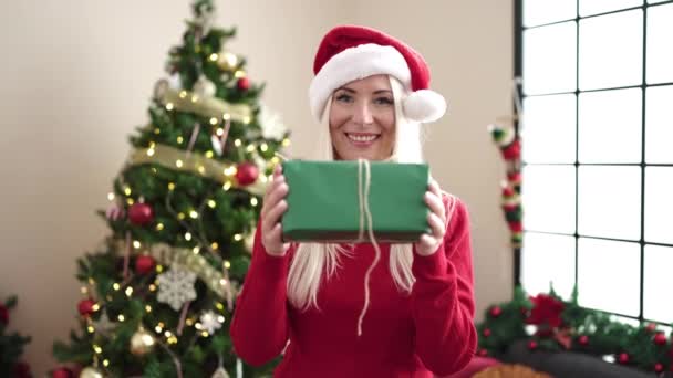 Νεαρή ξανθιά γυναίκα κρατώντας δώρο στέκεται δίπλα στο χριστουγεννιάτικο δέντρο στο σπίτι - Πλάνα, βίντεο