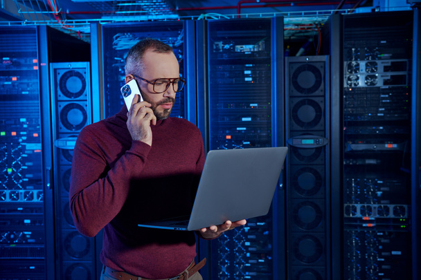 pensive data center specialist in turtleneck with glasses talking by phone and looking at his laptop - Photo, Image