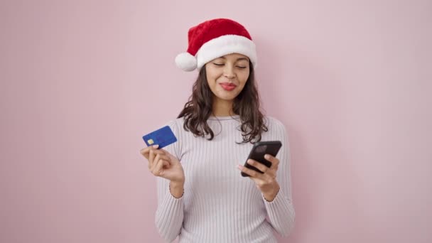 Молодая красивая латиноамериканка делает рождественские онлайн покупки со смартфоном на изолированном розовом фоне - Кадры, видео