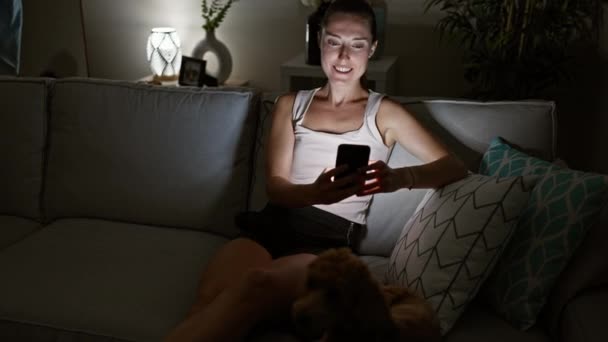 Mujer joven caucásica con perro usando teléfono inteligente sentado en un sofá en casa - Metraje, vídeo