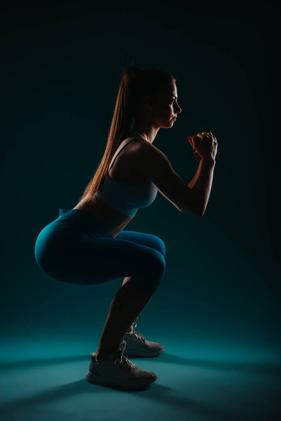 Una mujer confiada en un estudio oscuro hace ejercicio, mostrando sus músculos y la transformación de la aptitud. - Foto, imagen
