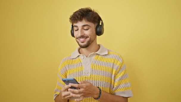 Cool arab férfi magabiztosan hallgat és táncol, hogy vidám zene az okostelefonján, létrehozva egy elszigetelt sárga háttér a boldogság és a pozitivitás - Felvétel, videó