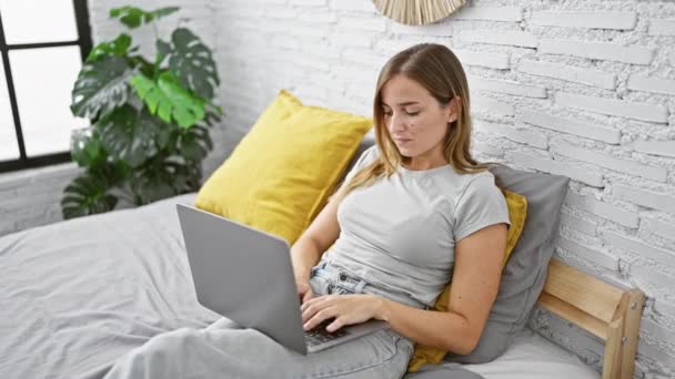 Молода блондинка використовує ноутбук, сидячи на ліжку в спальні
 - Кадри, відео