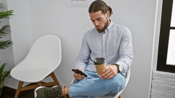 Joven hispano usando smartphone bebiendo café en la sala de espera - Metraje, vídeo