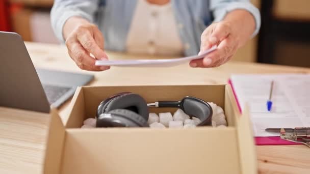 Middle age hispanic woman ecommerce business worker packing headphones on package at office - Footage, Video