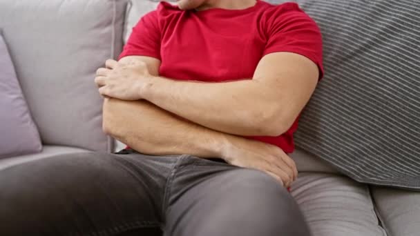 Ritratto di un giovane uomo ispanico bello seduto sul divano a casa, che gratta le braccia pruriginose - una possibile reazione allergica che causa irritazione e disagio alla pelle. - Filmati, video