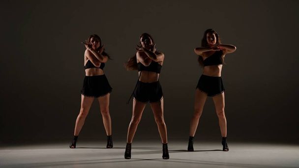 Group of women dancing heels dance in a studio. Plain shadowed background, spotlight. Black sexy costume, high heels. Modern sensual choreography. Full length. Promotional clip or advertisement. - Photo, image