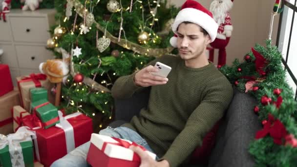 Fiatal spanyol férfi, hogy fotó ajándék okostelefonnal ül a kanapén karácsonyfa otthon - Felvétel, videó