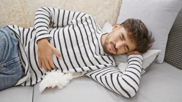 Fresco jovem adulto árabe homem, lidar com uma dor de estômago áspera, deitado preocupado em sua confortável sala de estar sofá em casa, tocando sua barriga magoada em agonia. - Filmagem, Vídeo