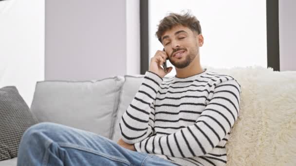 Hombre árabe joven agotado, sentado en su sofá de la sala de estar, absorto en una intensa conversación telefónica en su teléfono inteligente en casa - Metraje, vídeo