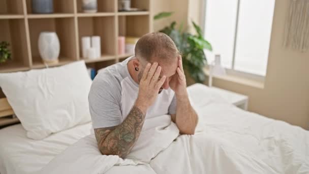 Vyčerpaný mladý muž, plný stresu, sedí na posteli, trpí bušící bolestí hlavy ve své klidné ložnici, odpočívá otřesený obavami. - Záběry, video