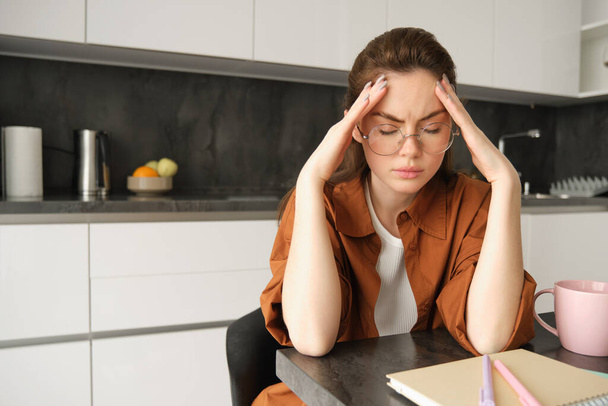 Портрет уставшей молодой женщины в очках, держащей руки за голову, страдающей болезненной мигренью, испытывающей боль в висках, сидящей с головной болью на кухне. - Фото, изображение