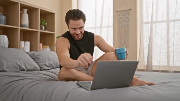 Joven hispano usando laptop bebiendo café sonriendo en el dormitorio - Metraje, vídeo