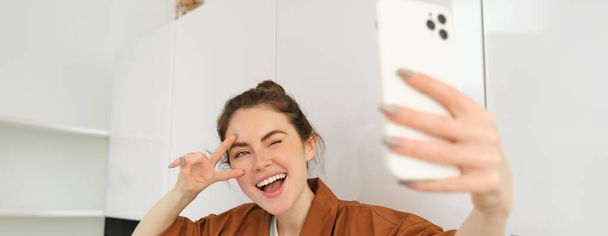 Close-up portret van positieve, gelukkige jonge vrouw selfie op mobiele telefoon, poseren voor foto op smartphone, tonen vrede teken en glimlachen naar de telefoon camera. - Foto, afbeelding