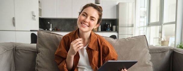 Portret van jonge brunette vrouw lezen op digitale tablet, tv-serie kijken op haar aanvraag, zitten op de bank in de woonkamer. - Foto, afbeelding