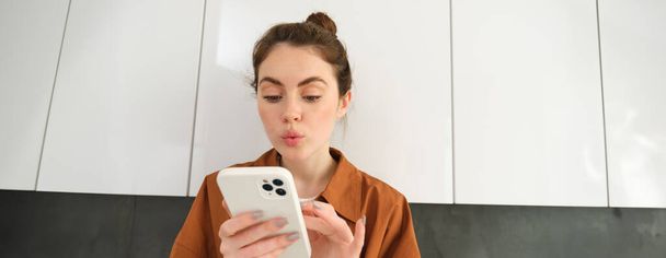 Fiatal nő portréja otthon, érdeklődéssel néz az okostelefon képernyőjére, az online vásárlás koncepciója, mobilalkalmazások vásárlása, telefonos alkalmazás használata. - Fotó, kép