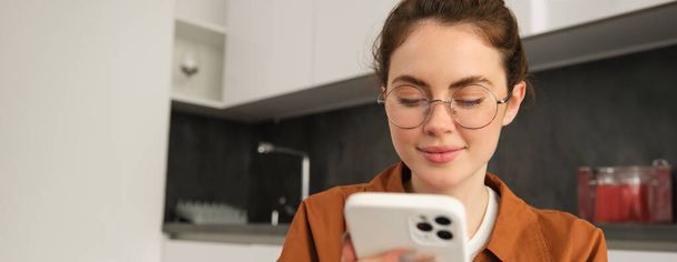 Πορτρέτο της νεαρής μελαχρινής γυναίκας με γυαλιά, κάθεται στο σπίτι με κινητό τηλέφωνο, χρησιμοποιεί το smartphone της και χαμογελά, διαβάζοντας το μήνυμα. - Φωτογραφία, εικόνα
