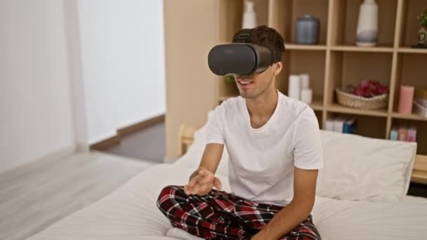 Sonriente joven jugador hispano disfrutando sentado en pijama en la cama del dormitorio, jugando videojuego usando gafas futuristas vr - Metraje, vídeo