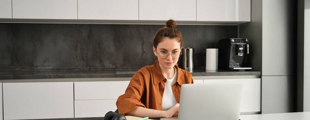 Concepto de equilibrio entre trabajo y vida. Mujer joven en gafas, trabajando desde casa, escribiendo en el ordenador portátil, estudiante haciendo la tarea en el ordenador, sentado en la cocina, freelancing. - Foto, imagen