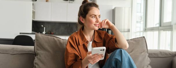 Πορτρέτο της κομψής σύγχρονης γυναίκας, θηλυκό μοντέλο κάθεται στο σπίτι στον καναπέ με smartphone, κοιτάζοντας έξω από το παράθυρο με τη σκέψη πρόσωπο, online ψώνια, μηνύματα στα μέσα κοινωνικής δικτύωσης. - Φωτογραφία, εικόνα