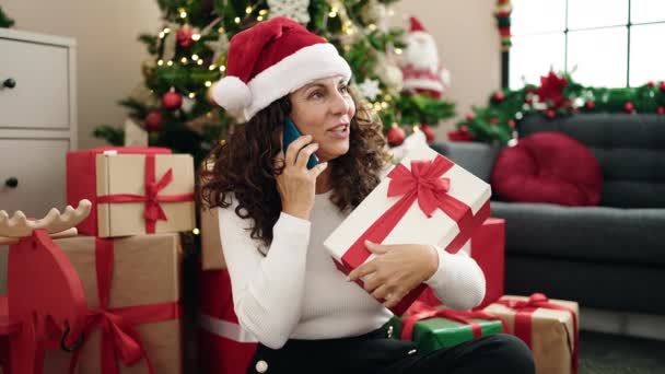 Hispanische Frau mittleren Alters spricht auf Smartphone und hält Weihnachtsgeschenk zu Hause - Filmmaterial, Video