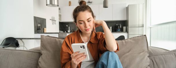 Jonge vrouw met rustig, ontspannen gezicht, lezen op smartphone, kijken naar mobiele telefoon scherm, zitten op de bank in de woonkamer. - Foto, afbeelding