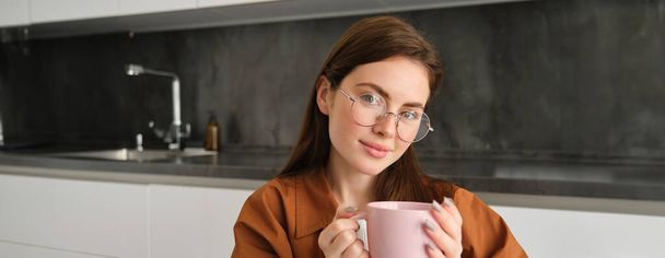 Ritratto di una giovane donna gioiosa che si gode una tazza di caffè a casa. Sorridente bella ragazza beve tè caldo in inverno. Donna eccitata che indossa occhiali e un maglione in una giornata autunnale. - Foto, immagini