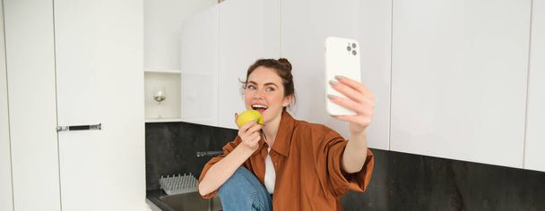 スマートフォンを使用してセルフィーを取る,カメラでリンゴを食べる,ソーシャルメディアアプリケーションのビデオを記録する,キッチンカウンターに座る. - 写真・画像