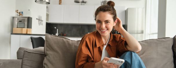 Όμορφο χαμογελαστό γυναικείο μοντέλο κάθεται στον καναπέ με το smartphone, κοιτάζοντας χαρούμενος στην κάμερα, κύλιση των μέσων κοινωνικής δικτύωσης, την αποστολή μηνυμάτων σε κάποιον, κάνοντας online ψώνια. - Φωτογραφία, εικόνα