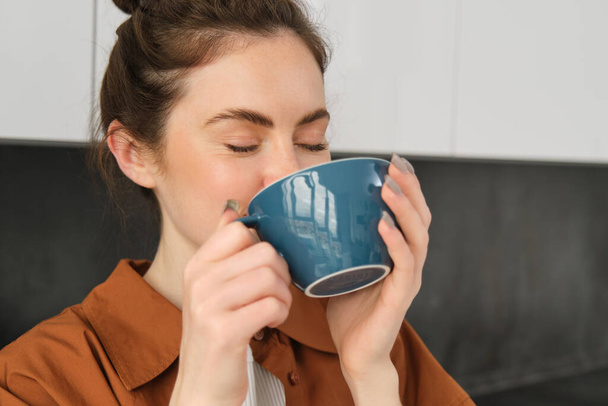 Ritratto di bella giovane donna che beve caffè, in piedi in cucina con grande tazza, gustando fagioli aromatici freschi, delizioso cappuccino. - Foto, immagini