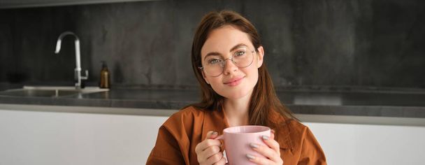 Ihastuttavan nuoren silmälasipäisen naisen muotokuva istuu kotona teekupin kanssa. Tyttö keittiössä, nauttii juomalla aromaattista kahvia. - Valokuva, kuva