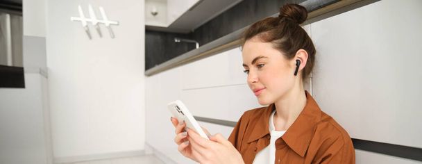 Close up portret młodej nowoczesnej kobiety siedzi na podłodze w kuchni, patrząc na jej smartfona, oglądając seriale wideo lub telewizyjne na streaming aplikacji usługowej, słuchając muzyki w słuchawkach bezprzewodowych. - Zdjęcie, obraz